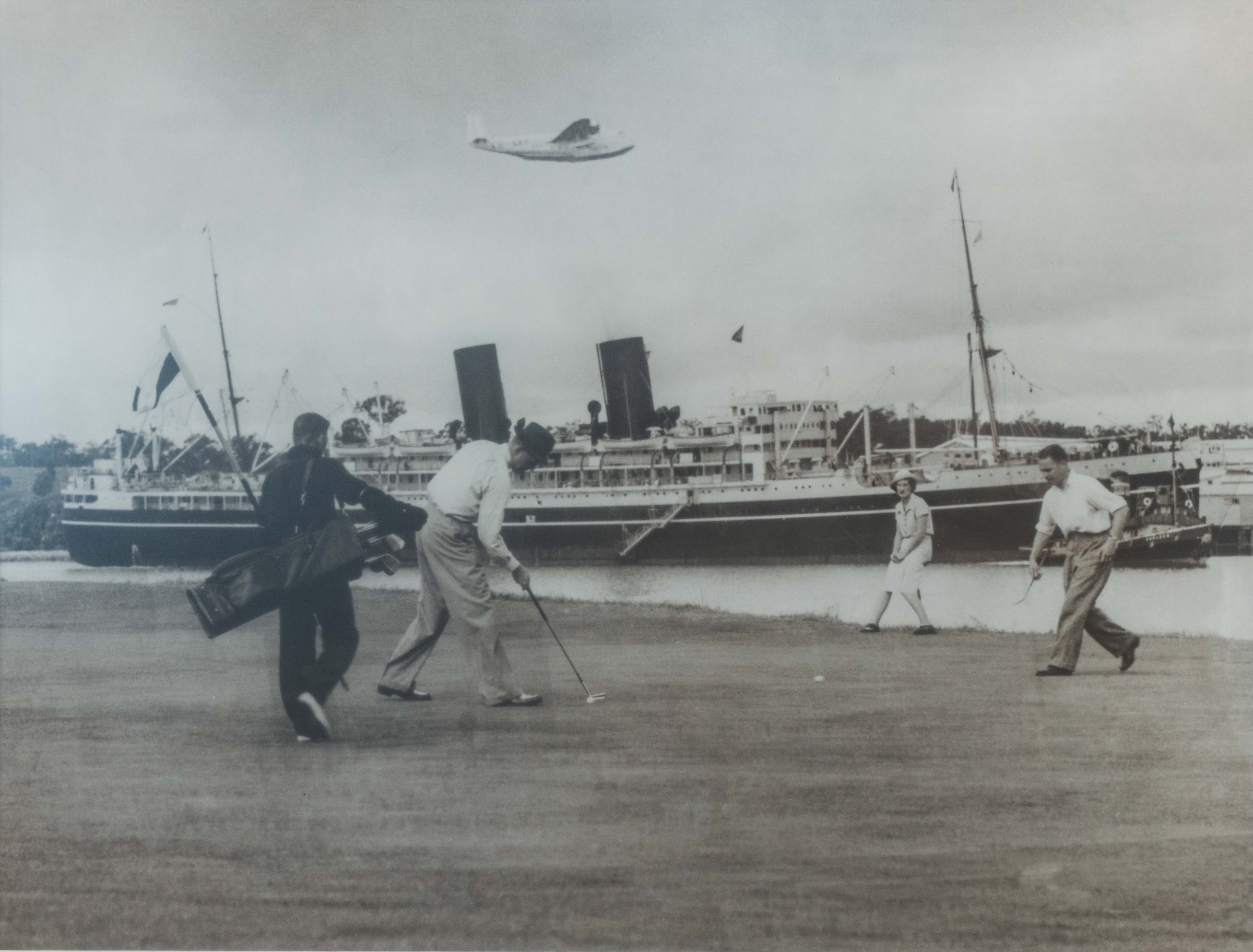 Golfers putting on a green at Royal Queensland in the 1930s, an ocean liner berthed in the Brisbane River directly behind them and a flying boat passing overhead